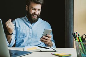 happy man holds a phone in his hand, sits at a desk in front of the computer